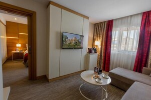 Habitación de hotel con una mesa redonda, un sofá, un televisor montado en la pared y un cuadro de un edificio.
