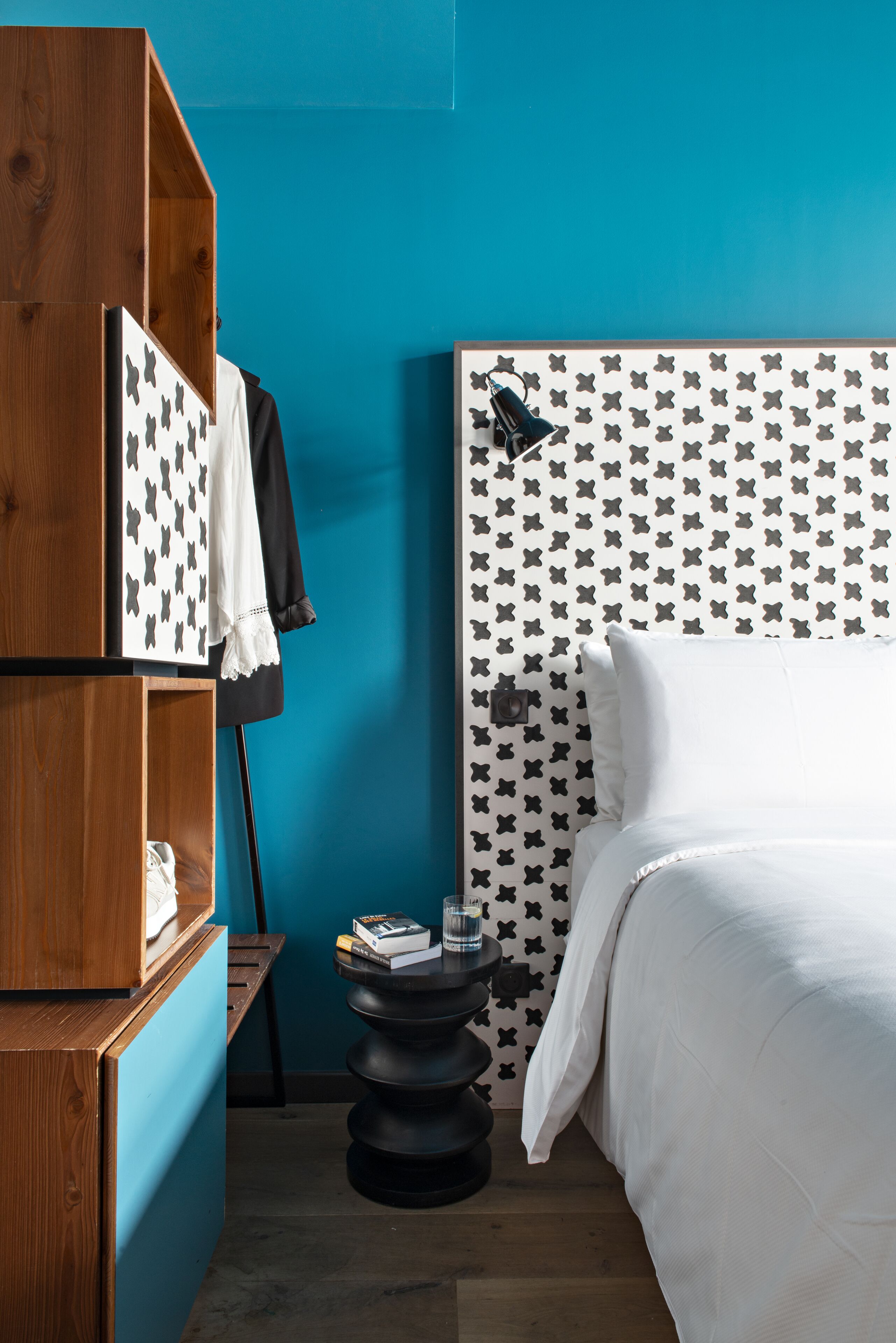 A bedroom with a wooden wardrobe, a bed with a patterned headboard, and a small round table with a book and a glass.