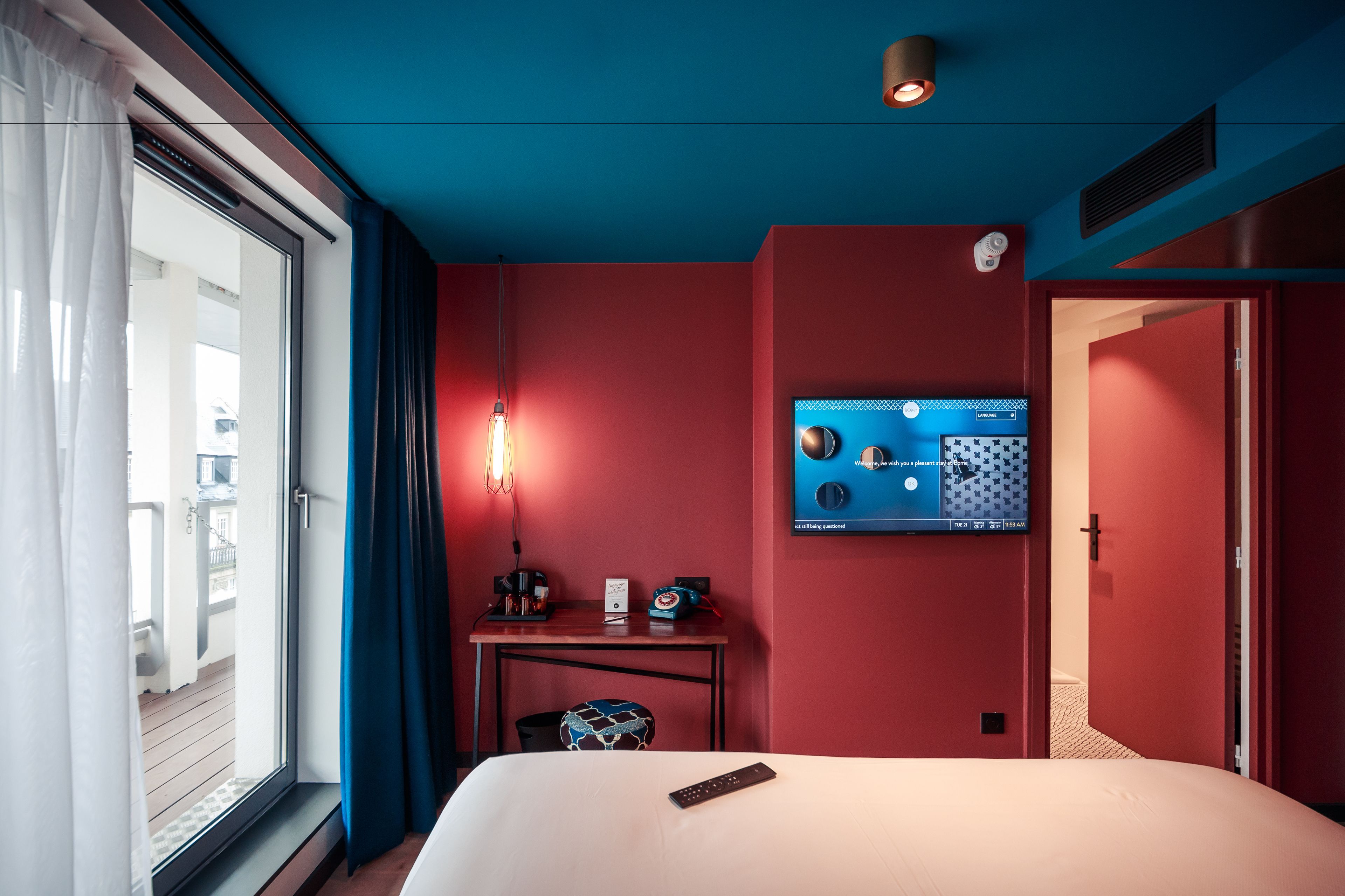 A hotel room with a bed, a desk with a lamp, a TV on the wall, and a window with curtains.