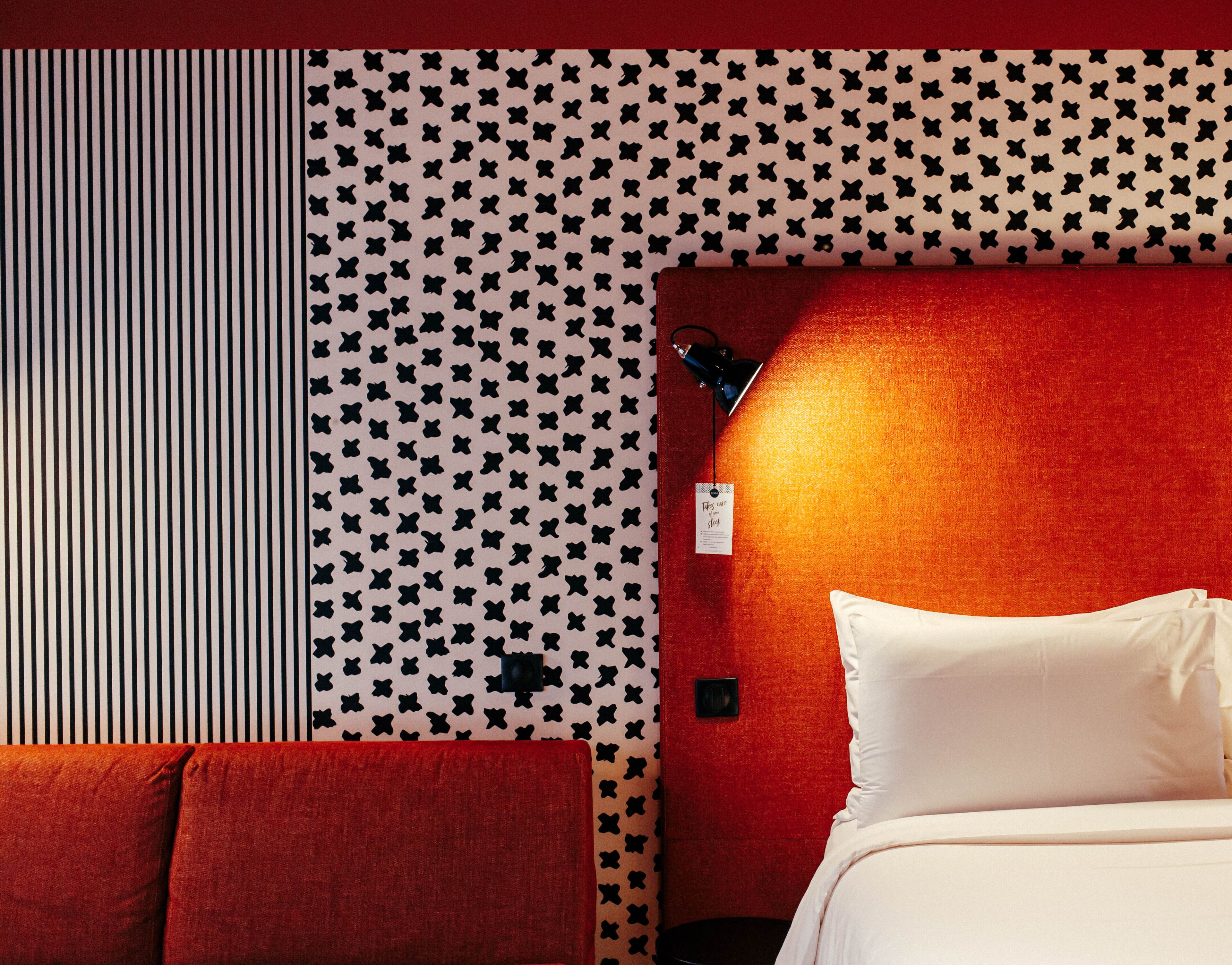 A hotel room with a striped wall, a patterned wall, and a red accent wall. There is a bed with white pillows and a red headboard.