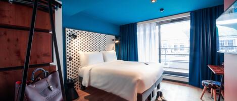A hotel room with a bed, a desk, a chair, a handbag, and a window with curtains.