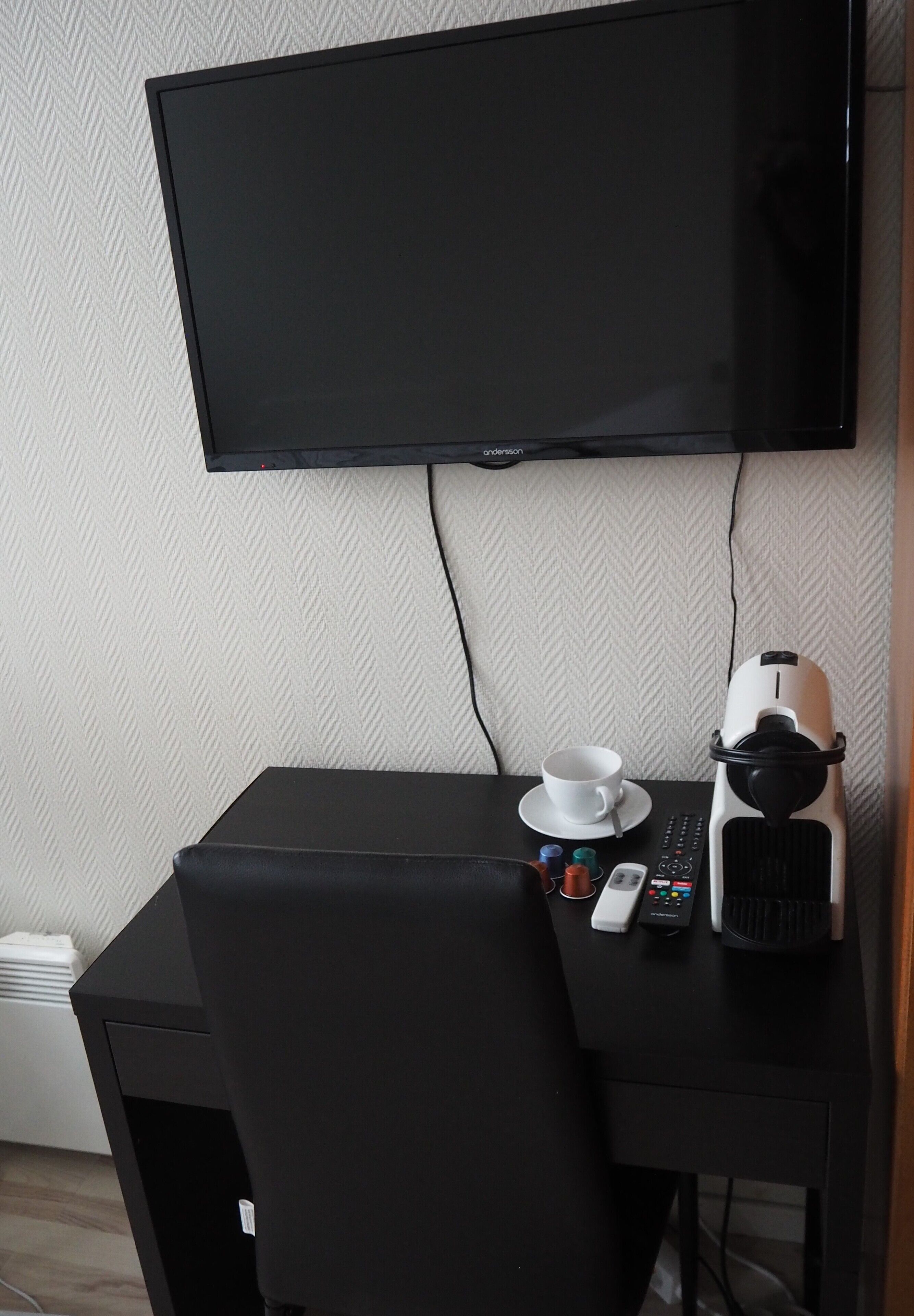 Un escritorio negro con un televisor montado encima, una taza blanca, un control remoto y una cafetera blanca.