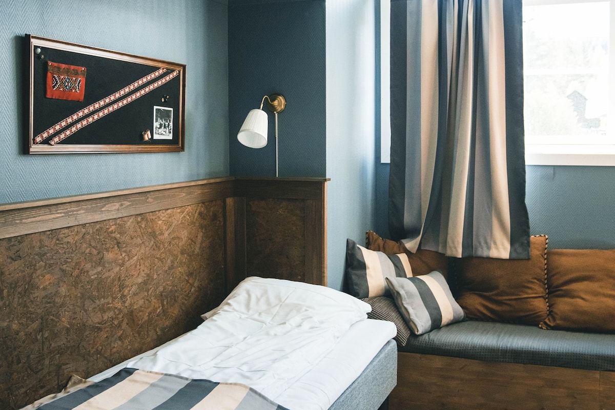 A bedroom with a bed, a wooden headboard, a wall-mounted lamp, a framed picture, and a window with curtains.