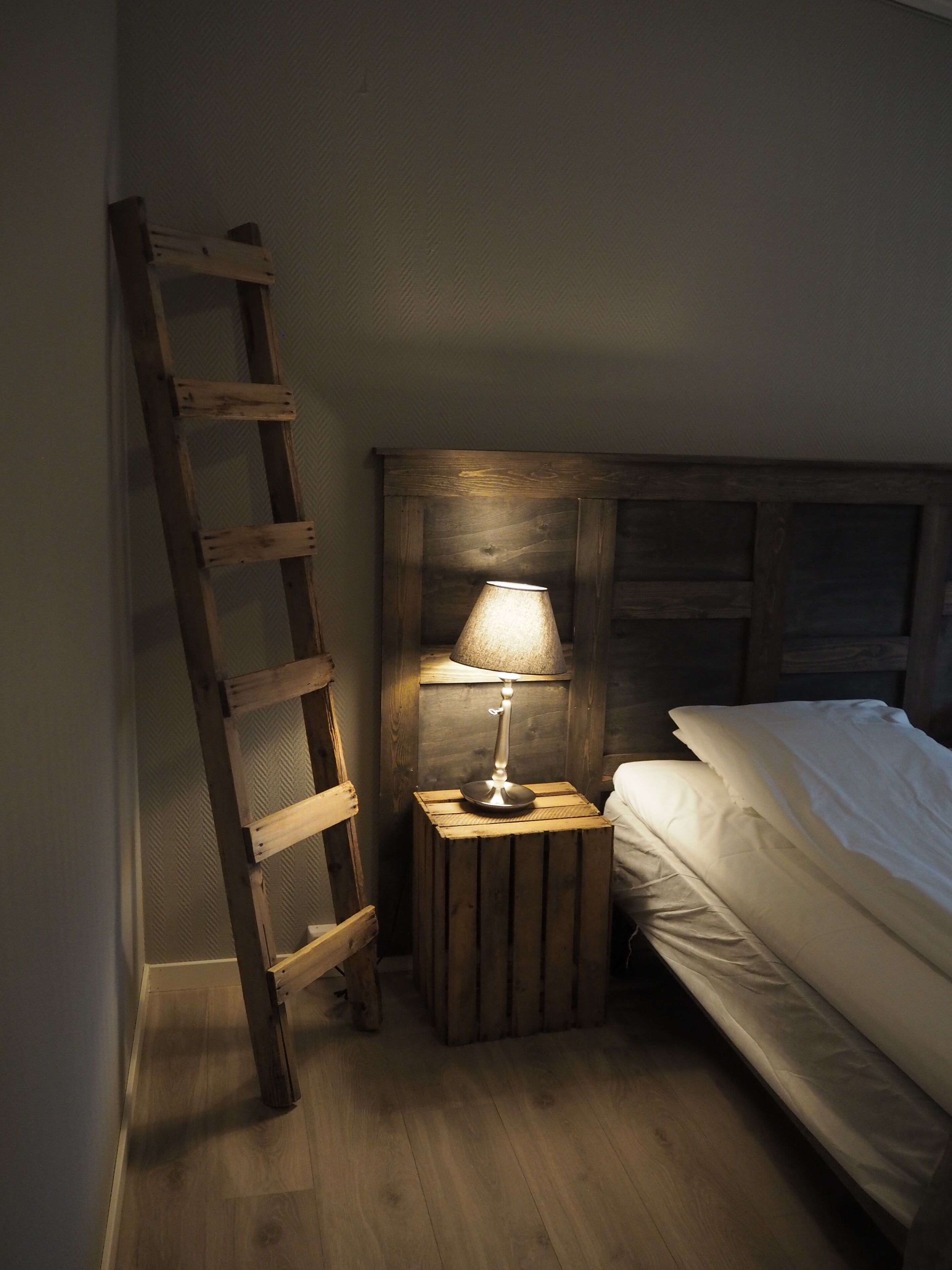 A wooden ladder leaning against a wall, a small wooden nightstand with a lamp, and a bed with white bedding.