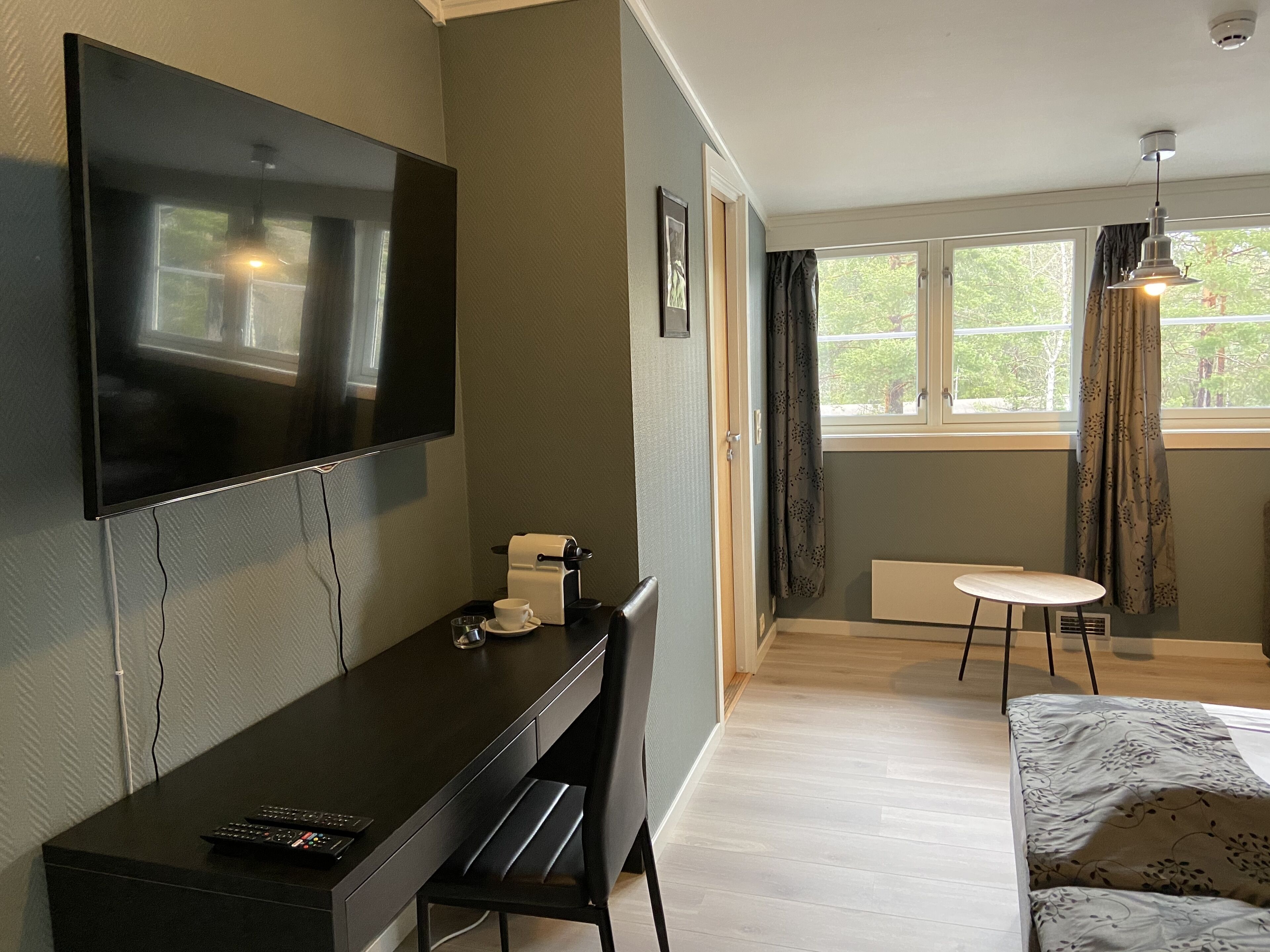 A hotel room with a flat-screen TV mounted on the wall, a desk with a coffee maker, a chair, a bed with a patterned bedspread, a small round table, and a window with curtains.