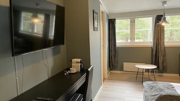 Habitación de hotel con televisor de pantalla plana montado en la pared, escritorio con cafetera, una silla, cama con colcha estampada, una mesita redonda y ventana con cortinas.