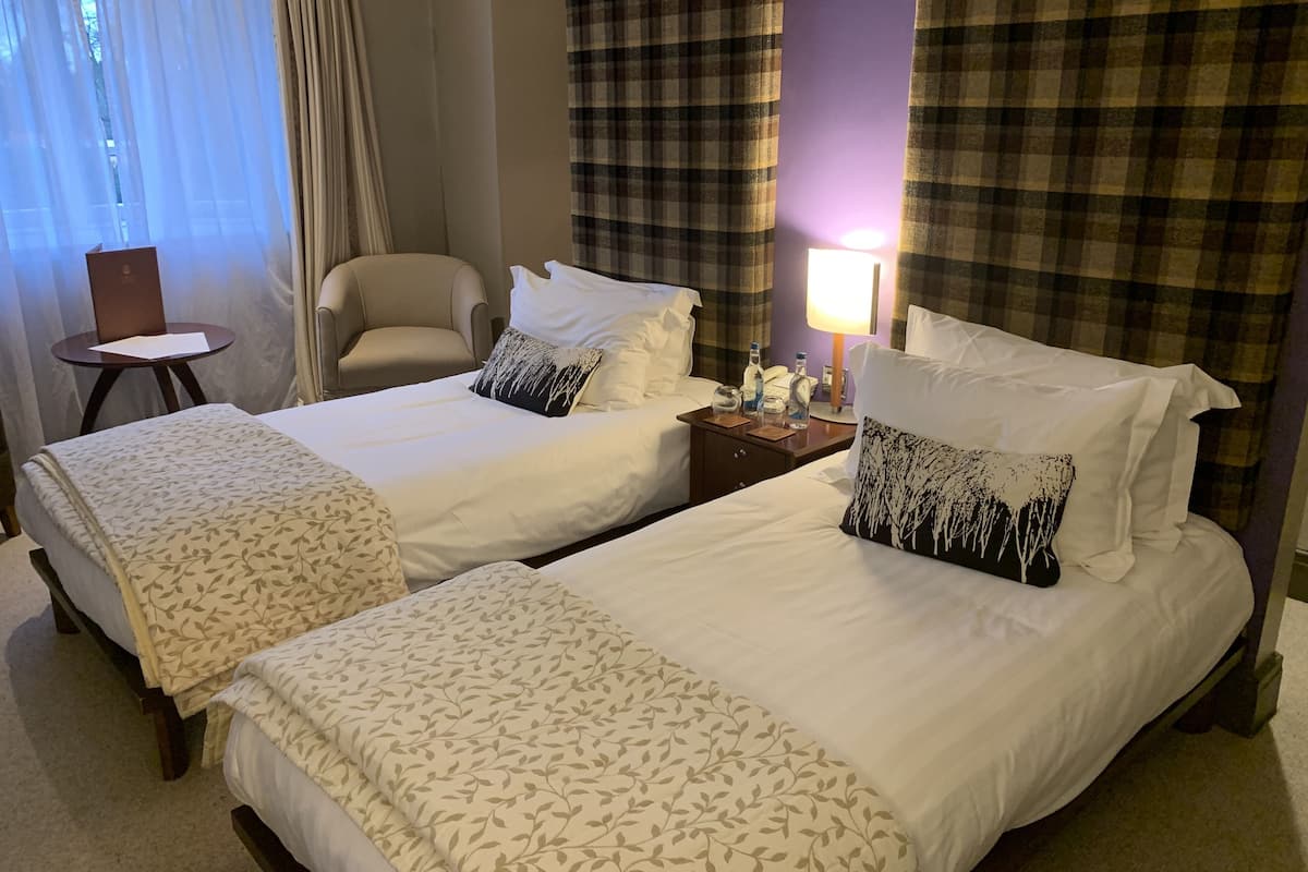 A hotel room with two beds, a chair, a small table, and a lamp.