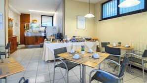 A dining area with tables set for breakfast, a kitchen in the background, and a large window with blinds. - Hotel Anna Siena Nord (Monteriggioni)