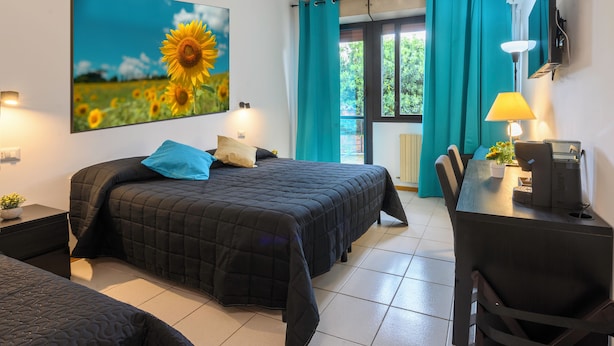 A bedroom with a bed, a desk, and a chair. There is a window with curtains and a painting of sunflowers on the wall. - Hotel Anna Siena Nord (Monteriggioni)