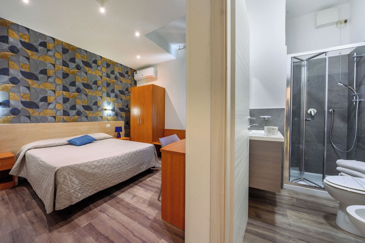 Un dormitorio moderno con cama, armario de madera, escritorio y baño con ducha e inodoro.