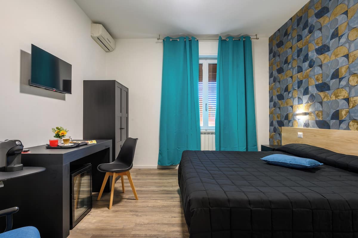 Una habitación de hotel moderna con una cama, un escritorio con computadora, un televisor y una ventana con cortinas.