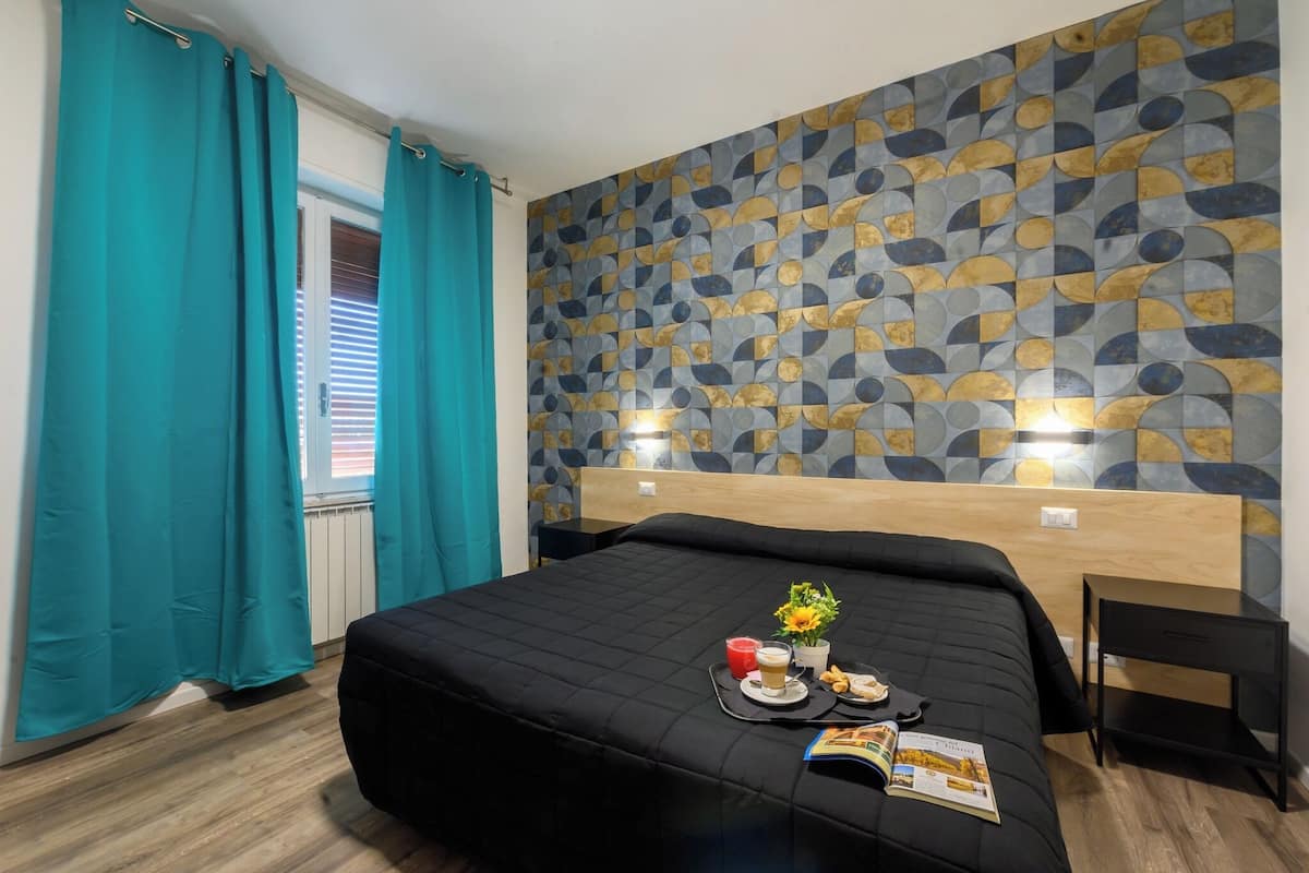 Habitación de hotel con cama, cortinas azul verdosas, pared con diseño geométrico y una pequeña mesa con bebidas y aperitivos.