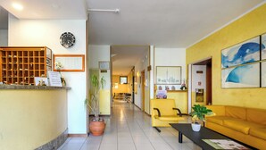 A hotel lobby with a reception desk, a yellow sofa, a coffee table, and a potted plant. - Hotel Anna Siena Nord (Monteriggioni)