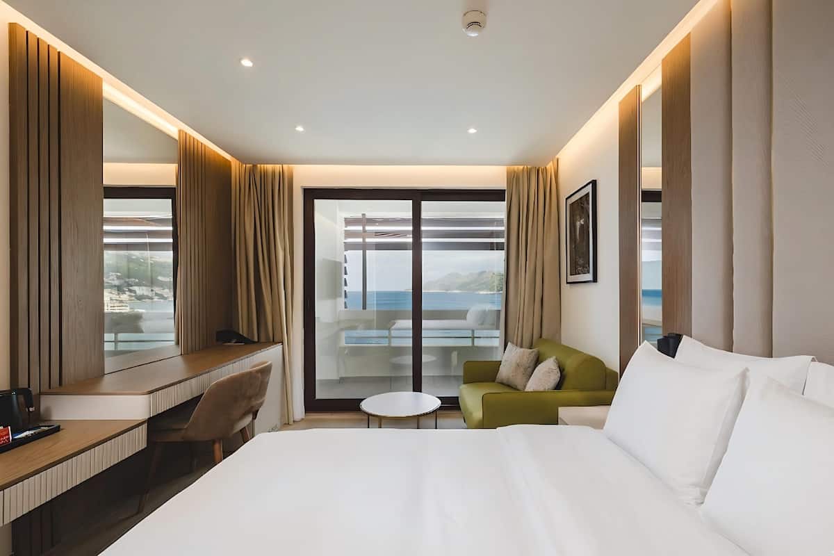 Une chambre d’hôtel avec un grand lit, un bureau, une chaise, une petite table, un canapé et une grande fenêtre donnant sur la mer.