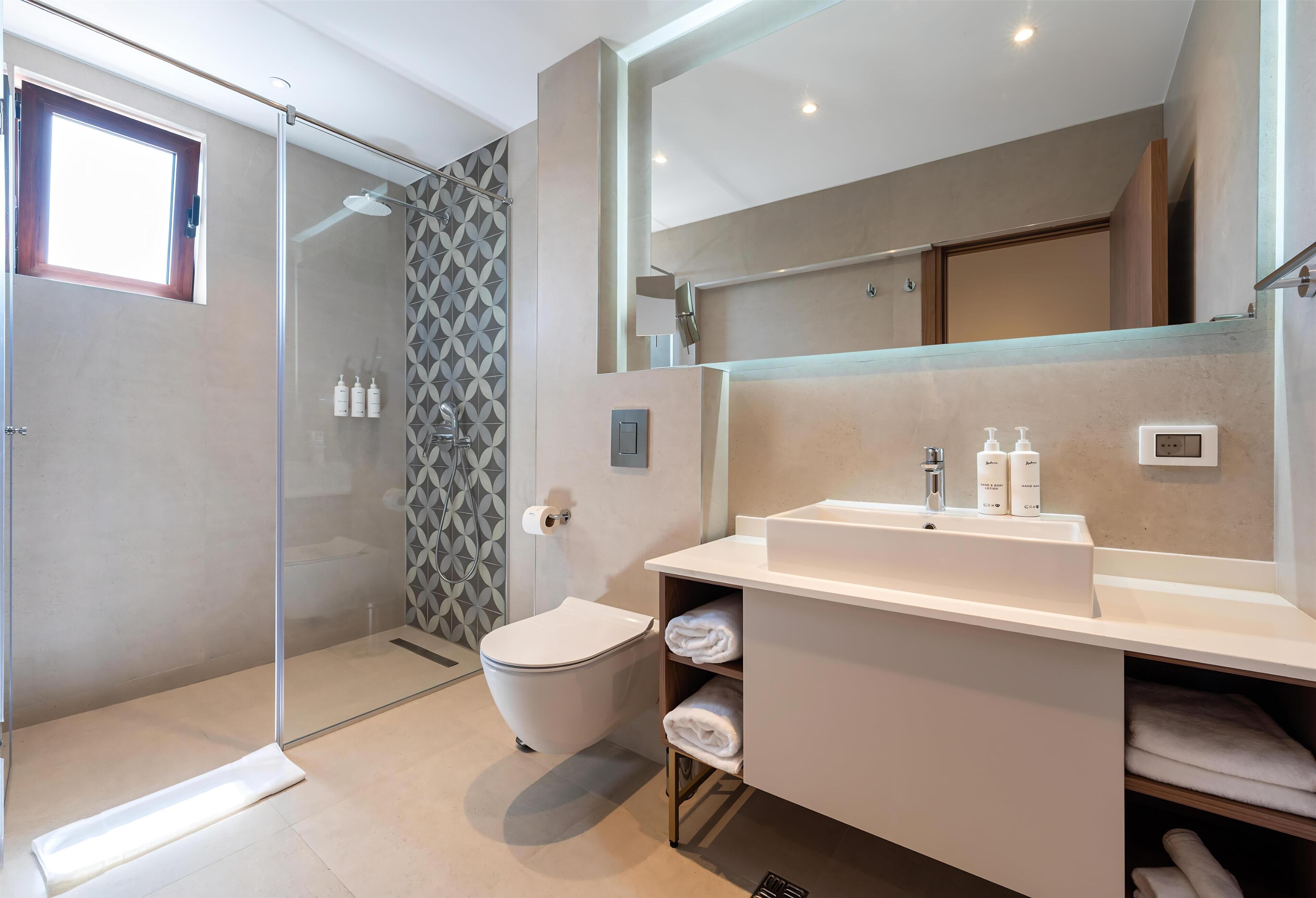 Et moderne badeværelse med et glasbadeværelse, toilet, håndvask med toiletartikler og et stort spejl.