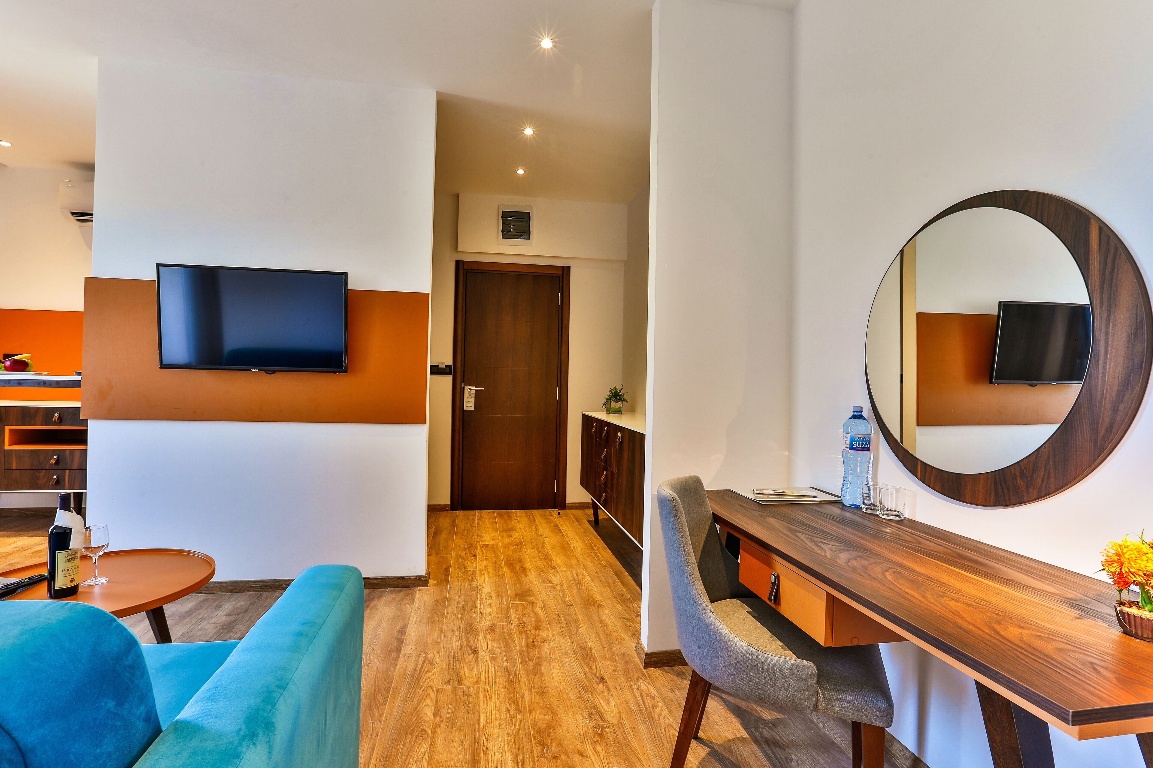 Une chambre d’hôtel moderne dotée d’un téléviseur à écran plat fixé au mur, d’un bureau en bois avec un miroir, d’un canapé bleu et d’un plancher en bois.