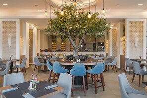Ein modernes Restaurant mit einem zentralen Baum, runden Tischen und blauen Stühlen.