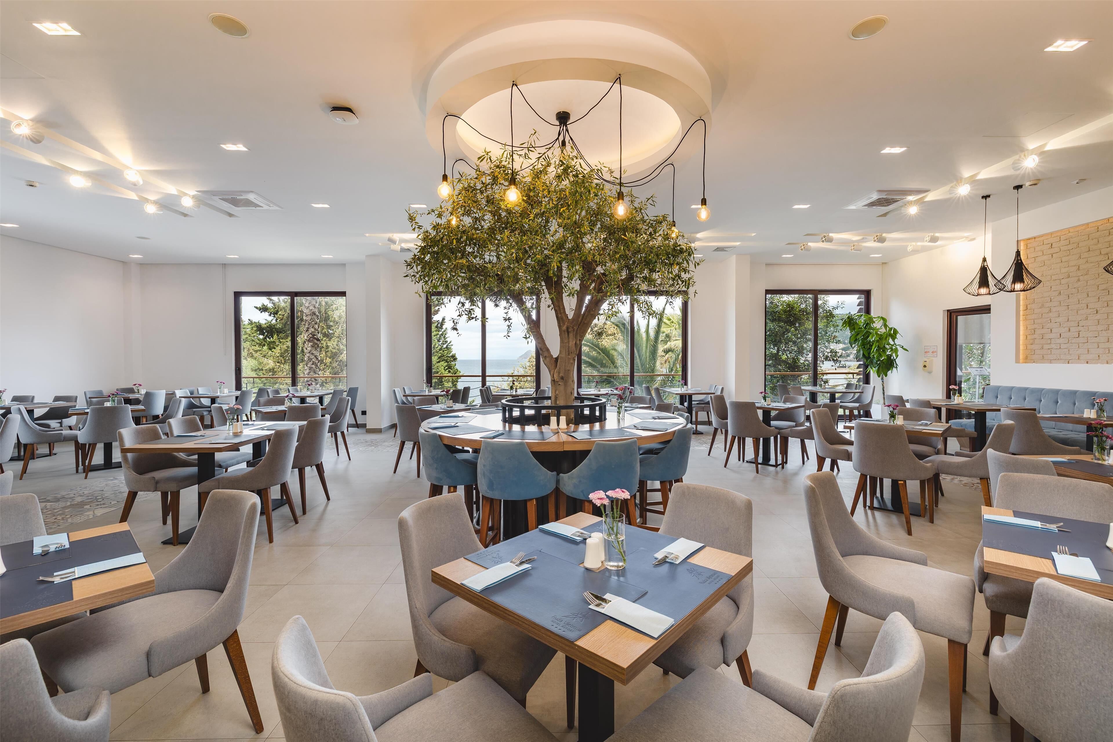 Un comedor amplio con mesas redondas y sillas modernas, un gran árbol como centro de mesa y ventanas grandes que dejan entrar luz natural.