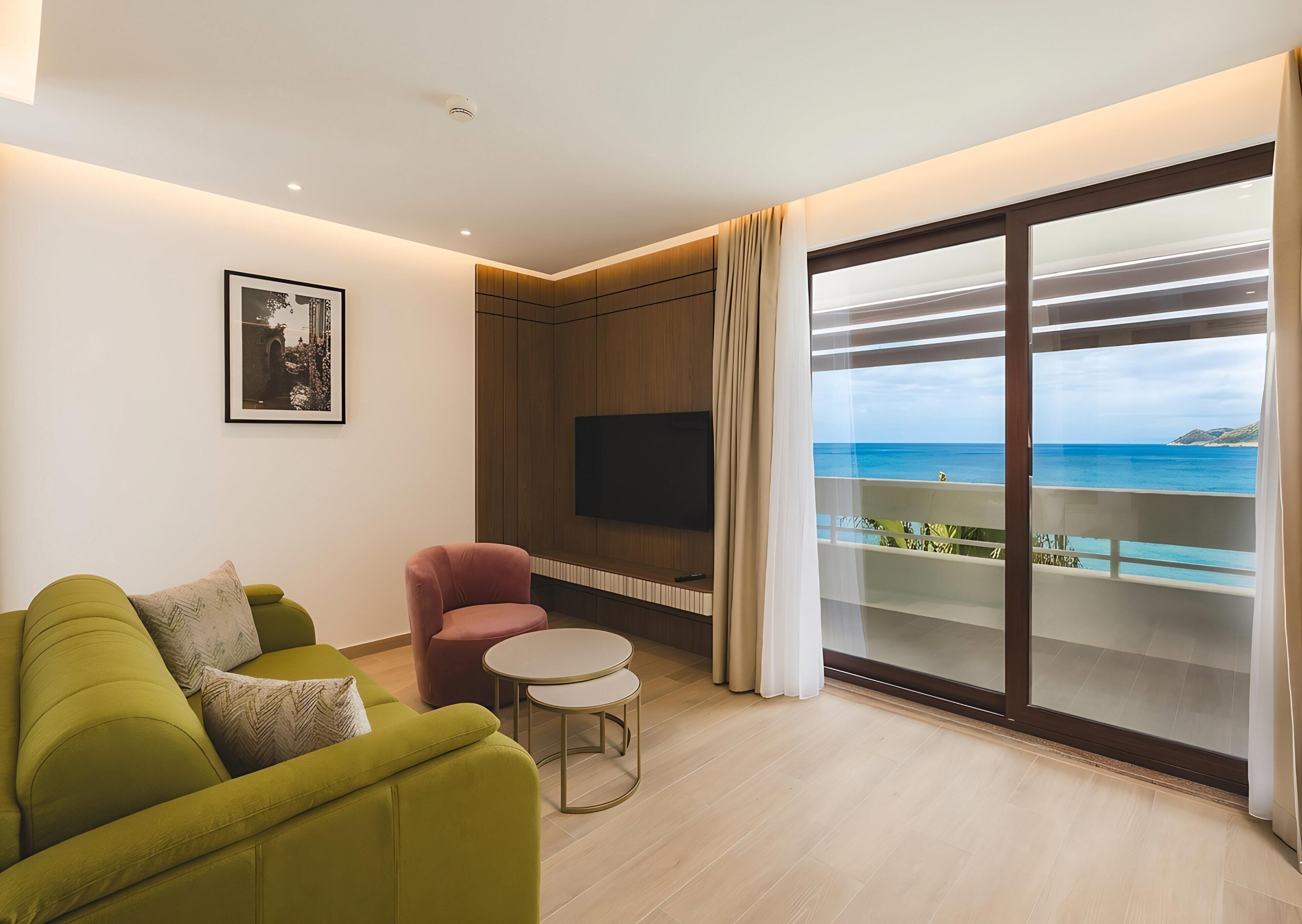 A modern living room with a green sofa, a red armchair, a small round table, and a TV mounted on a wooden panel wall. A large sliding glass door leads to a balcony with a view of the sea.
