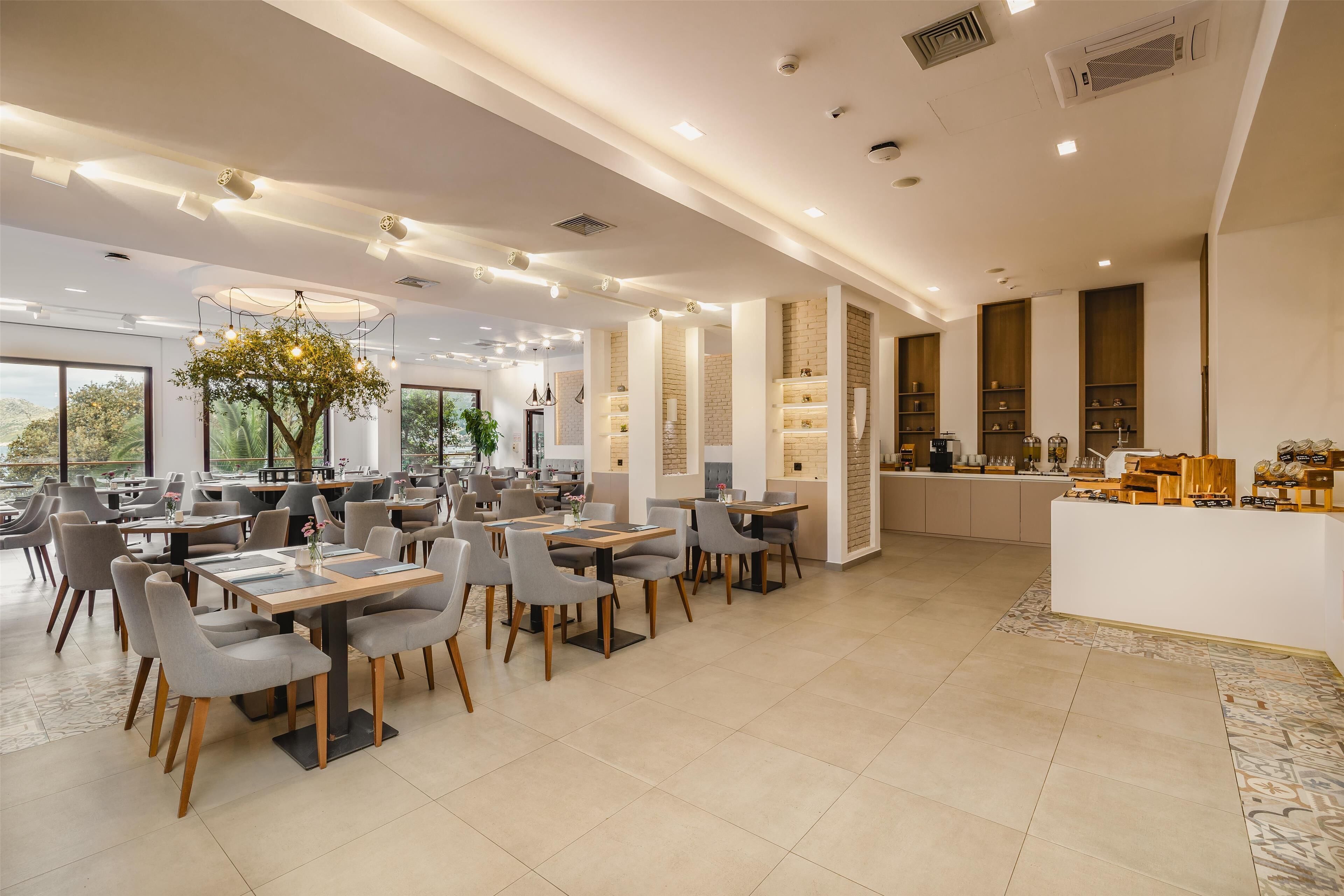 Una sala da pranzo moderna con diversi tavoli e sedie, un lampadario centrale e una zona cucina con scaffali ed elettrodomestici.