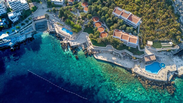 An aerial view of a coastal resort with clear waters, a swimming pool, and multiple buildings with red-tiled roofs. - Radisson Resort Ruza Vjetrova (Bar)