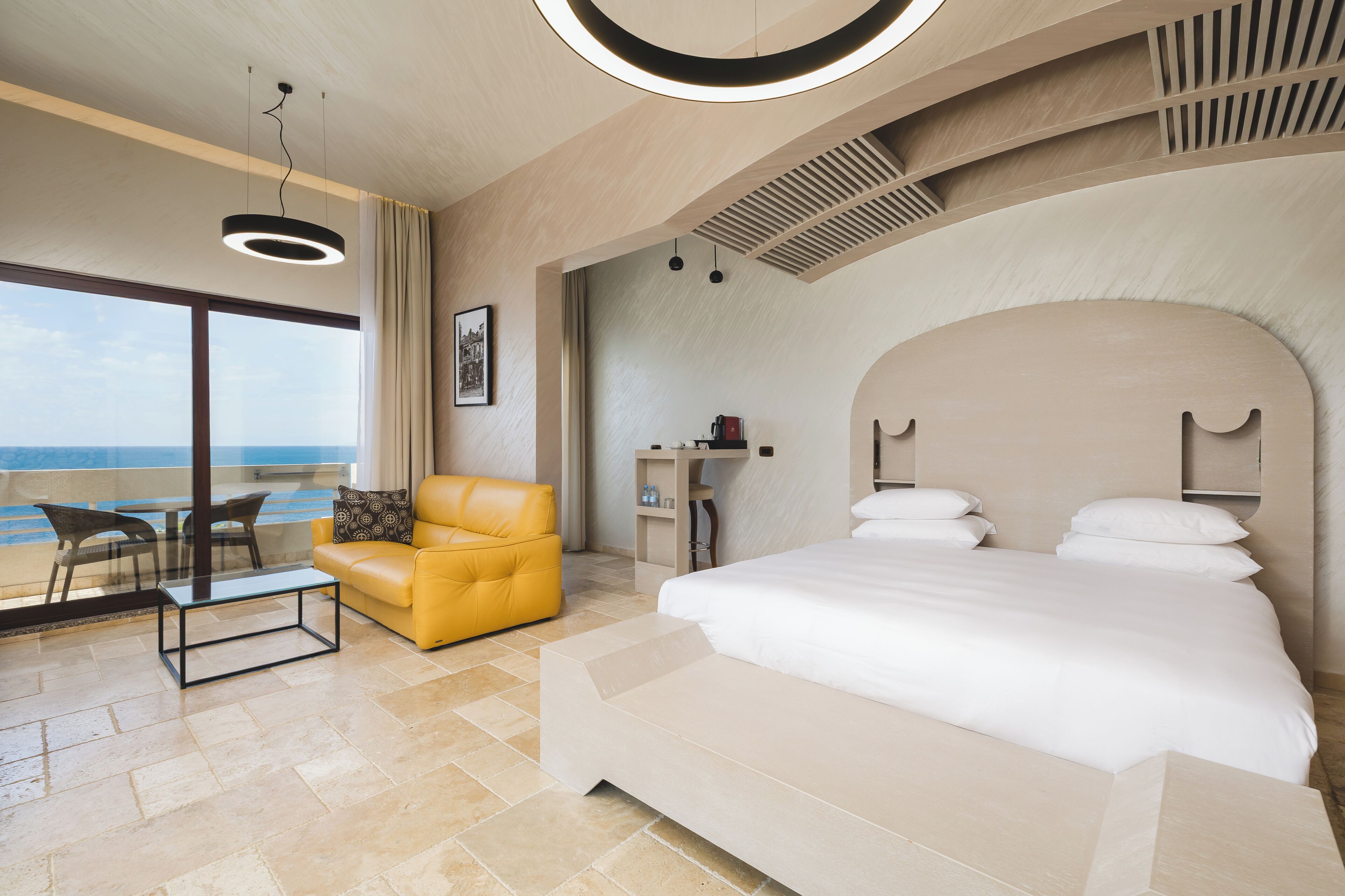 A modern hotel room with a large bed, a sofa, a desk, and a view of the sea.