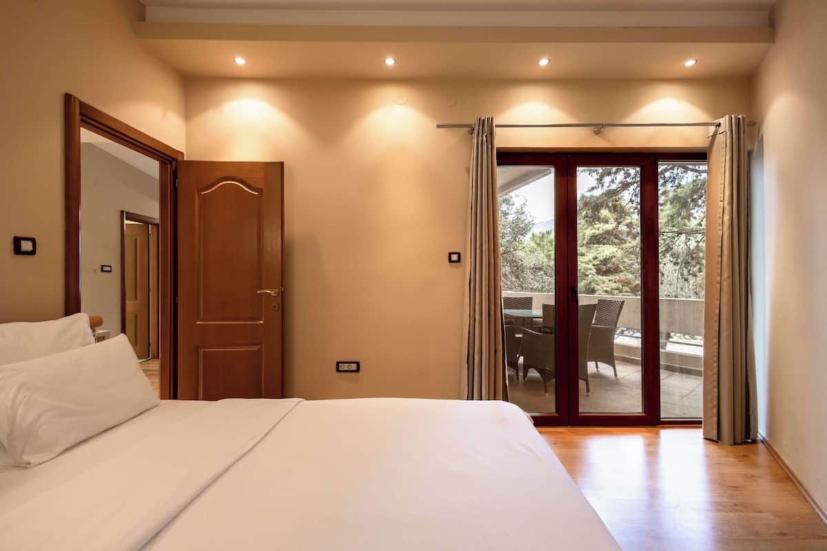 Une chambre avec un grand lit, une porte en bois et une porte-fenêtre coulissante donnant sur une terrasse extérieure aménagée avec du mobilier.