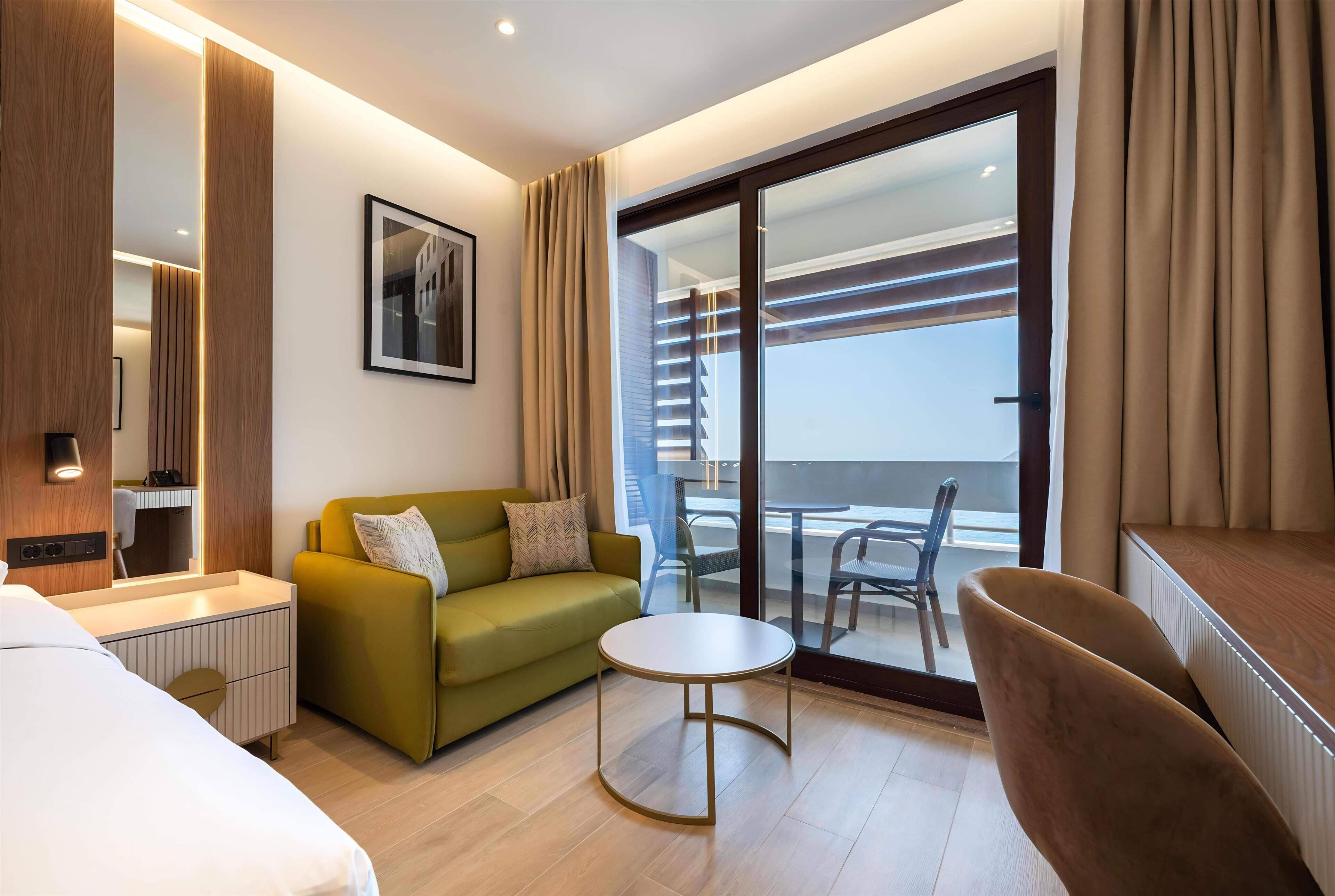 A hotel room with a bed, a green sofa, a round table, a chair, a balcony with a view, and a large window.