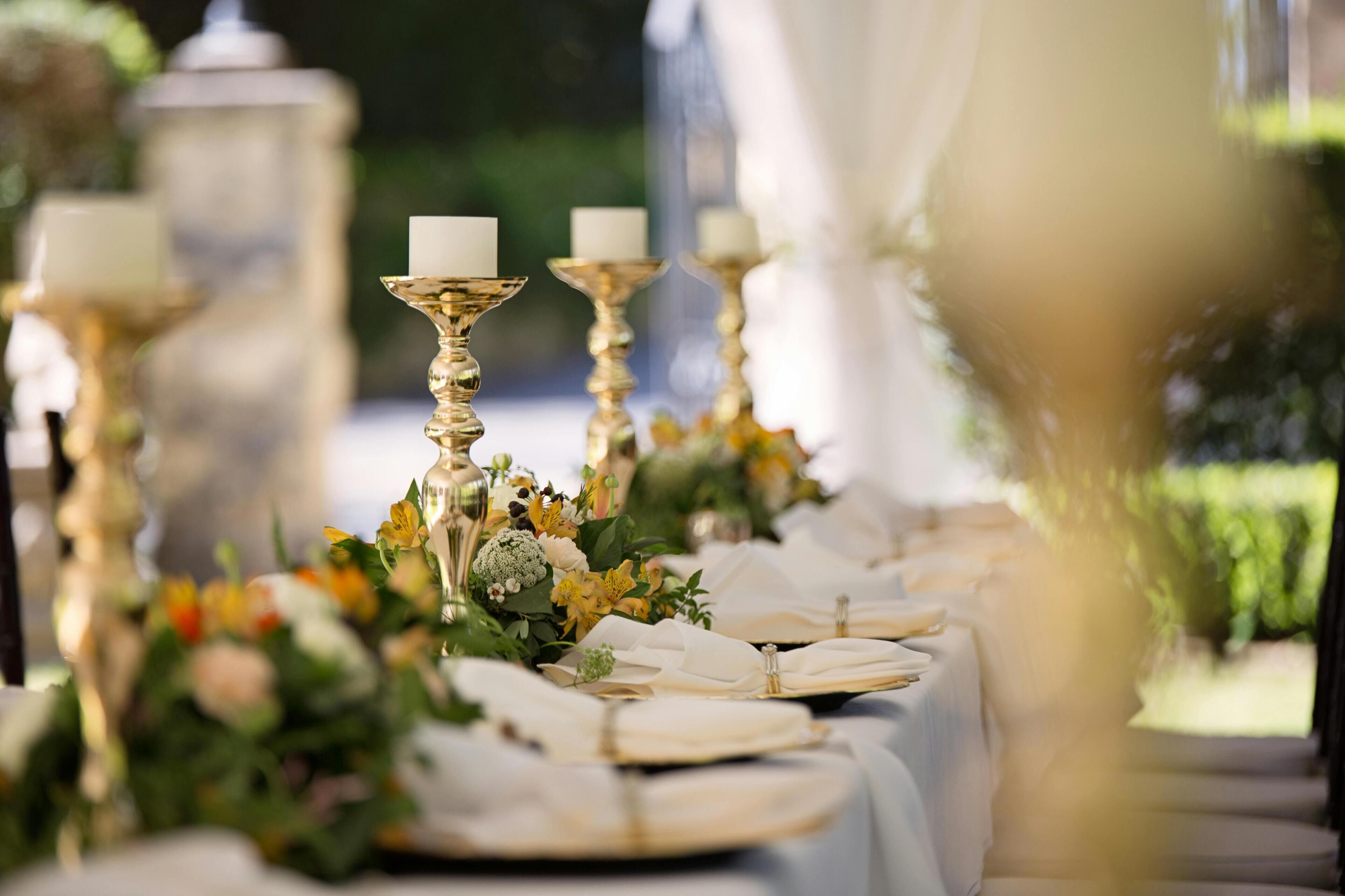 Una mesa dispuesta con servilletas blancas, candelabros dorados y centros de mesa florales.