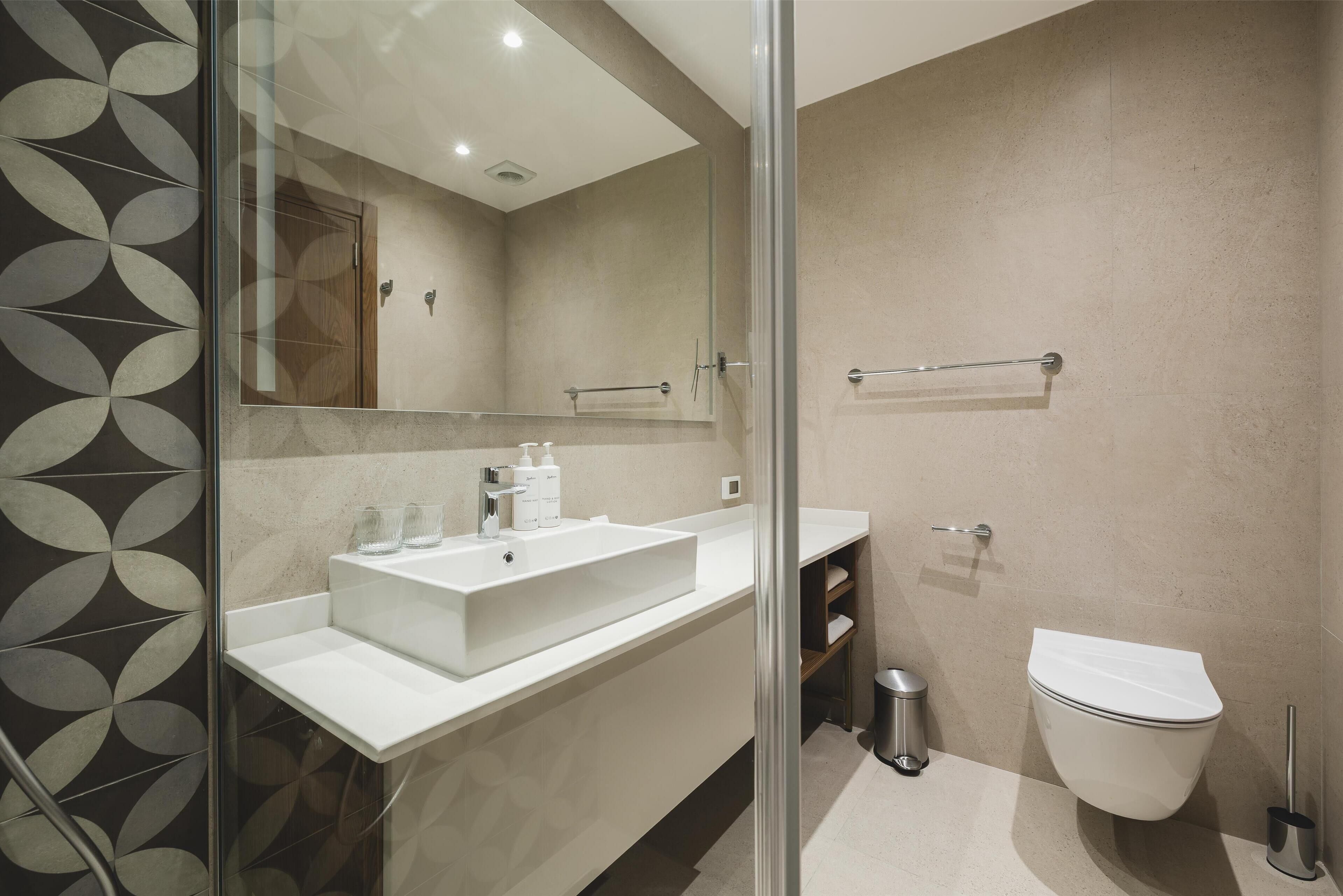 Un baño moderno con un espejo grande, un lavamanos blanco, un inodoro y una zona de ducha con mampara de vidrio.