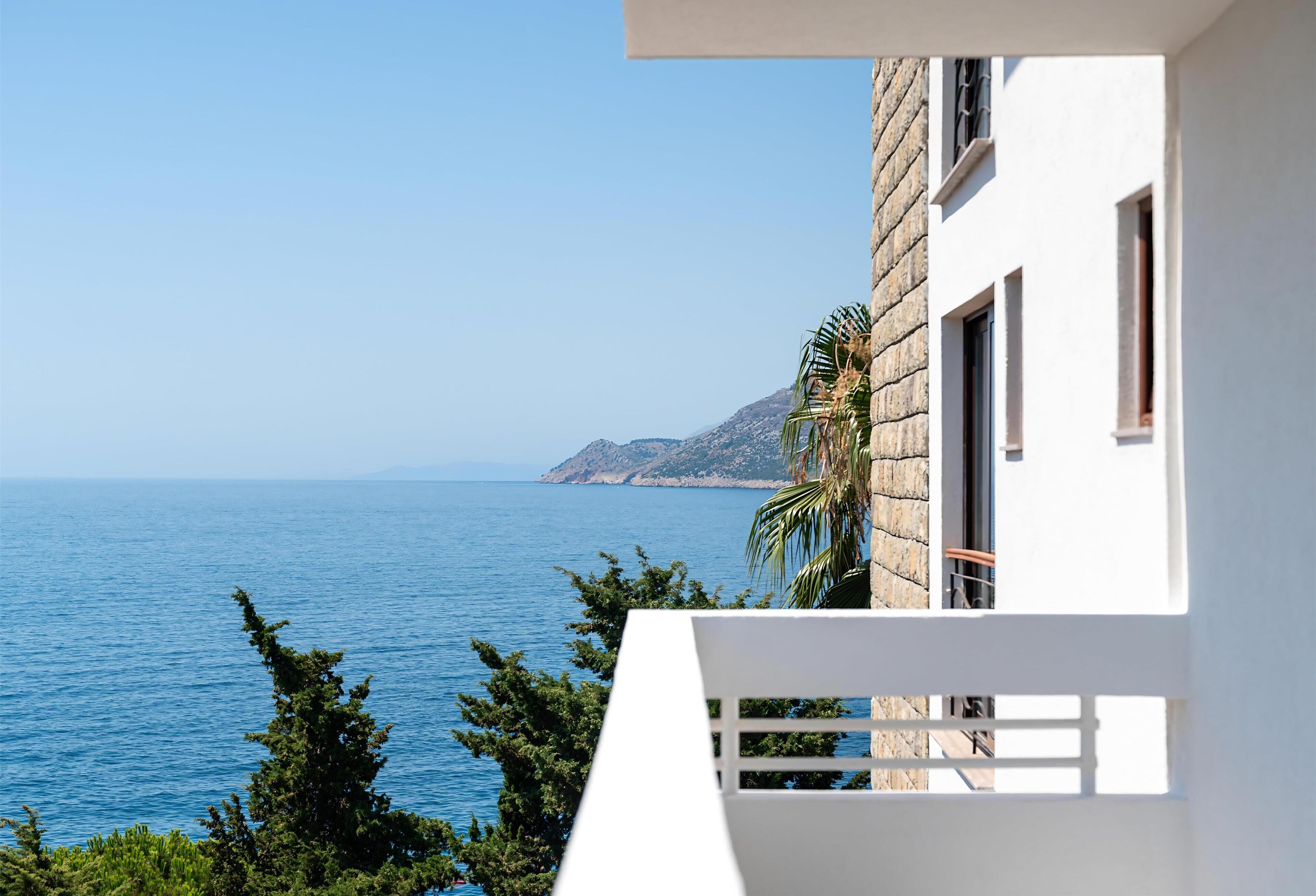Een wit gebouw met een balkon met uitzicht op een heldere blauwe zee en verre bergen.