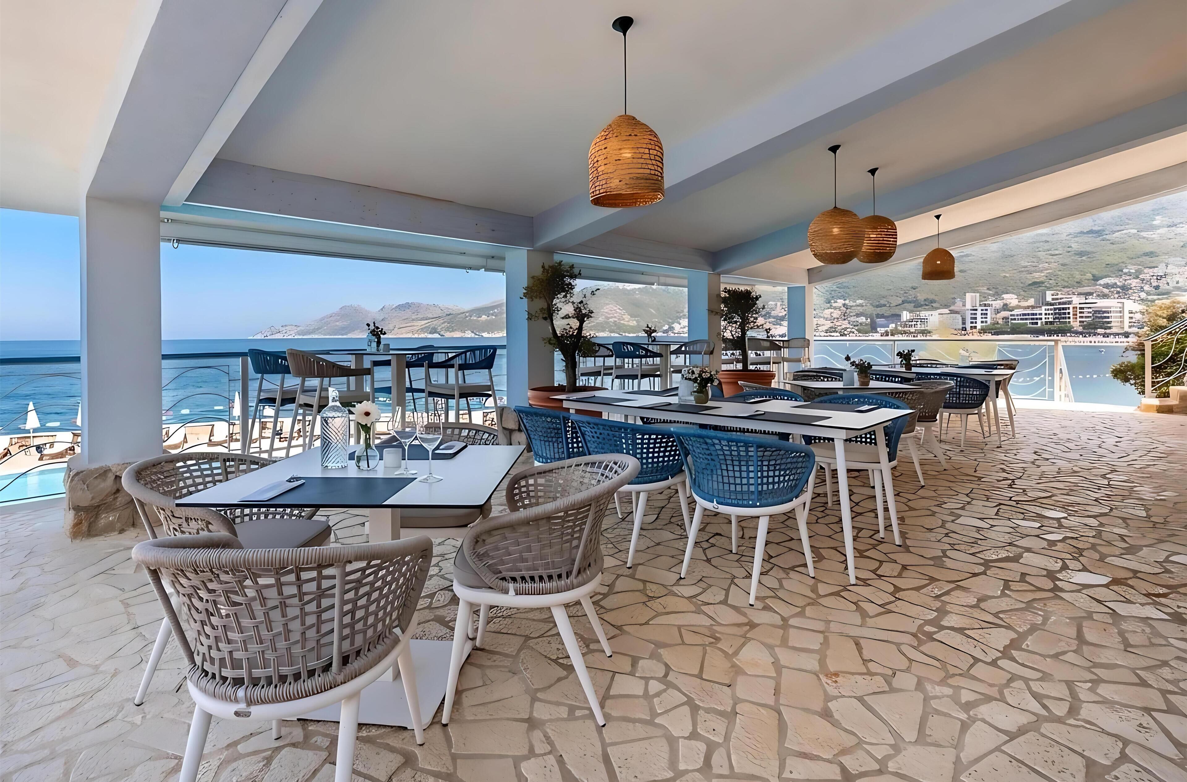 An outdoor dining area with tables and chairs, a stone floor, and a view of the sea and mountains.