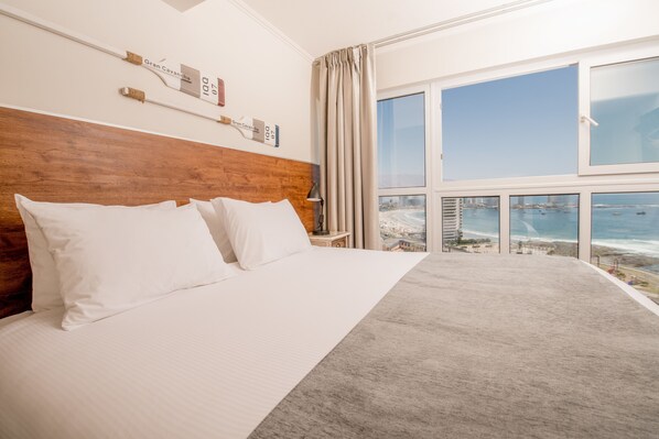 Un cuarto de hotel con una cama grande, un cabecero de madera, vista al océano y un paisaje urbano a través de las ventanas.