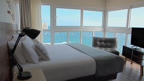 Habitación de hotel con una cama grande, un escritorio con lámpara, una silla y televisor. La habitación tiene ventanas amplias con vista al océano y edificios.