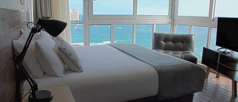 A hotel room with a large bed, a desk with a lamp, a chair, and a TV. The room has large windows with a view of the ocean and buildings.