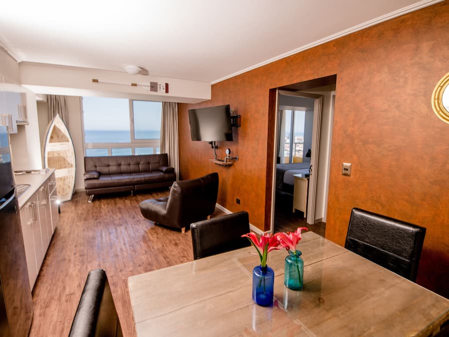 A modern hotel room with a dining area, a sofa, a TV, and a view of the sea.