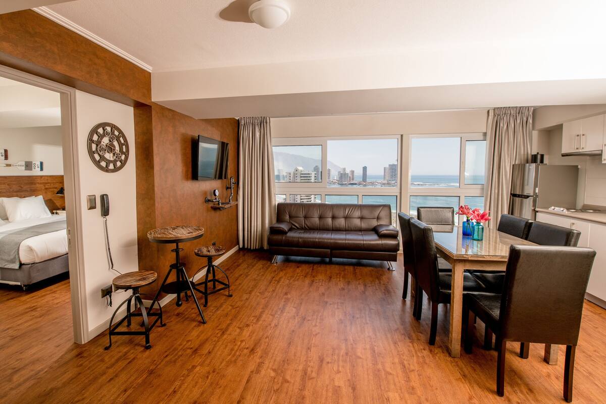 Una habitación de hotel moderna con suelo de madera, un sofá, una mesa de comedor con sillas, un televisor montado en la pared y vistas a la ciudad a través de amplios ventanales.