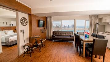A modern hotel room with a wooden floor, a sofa, a dining table with chairs, a TV mounted on the wall, and a city view through large windows.