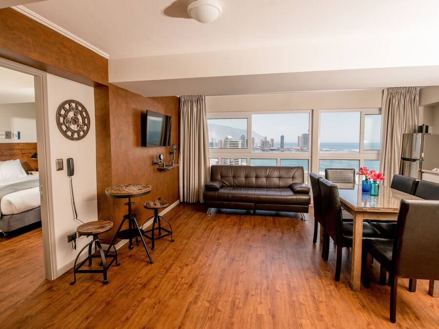 A modern hotel room with a wooden floor, a sofa, a dining table with chairs, a TV mounted on the wall, and a city view through large windows.