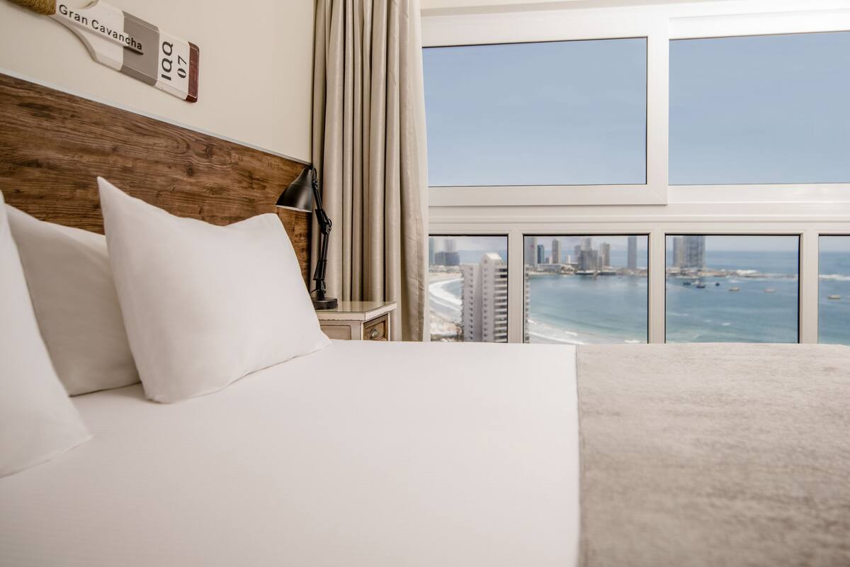 Habitación de hotel con una cama grande, una mesita de noche, una lámpara y vistas al océano y al horizonte urbano a través de amplios ventanales.