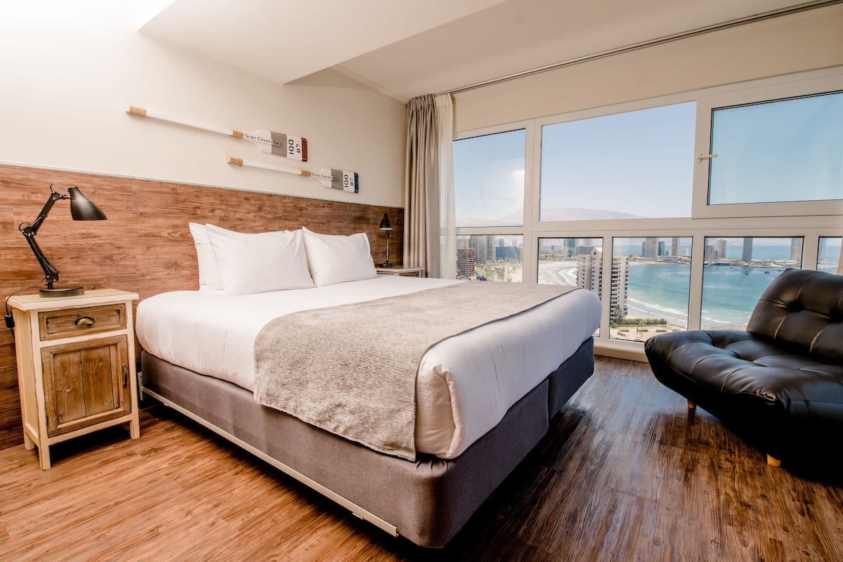 Habitación de hotel moderna con una cama grande, una mesita de noche, un sillón de cuero y vistas a la ciudad y al océano a través de amplios ventanales.