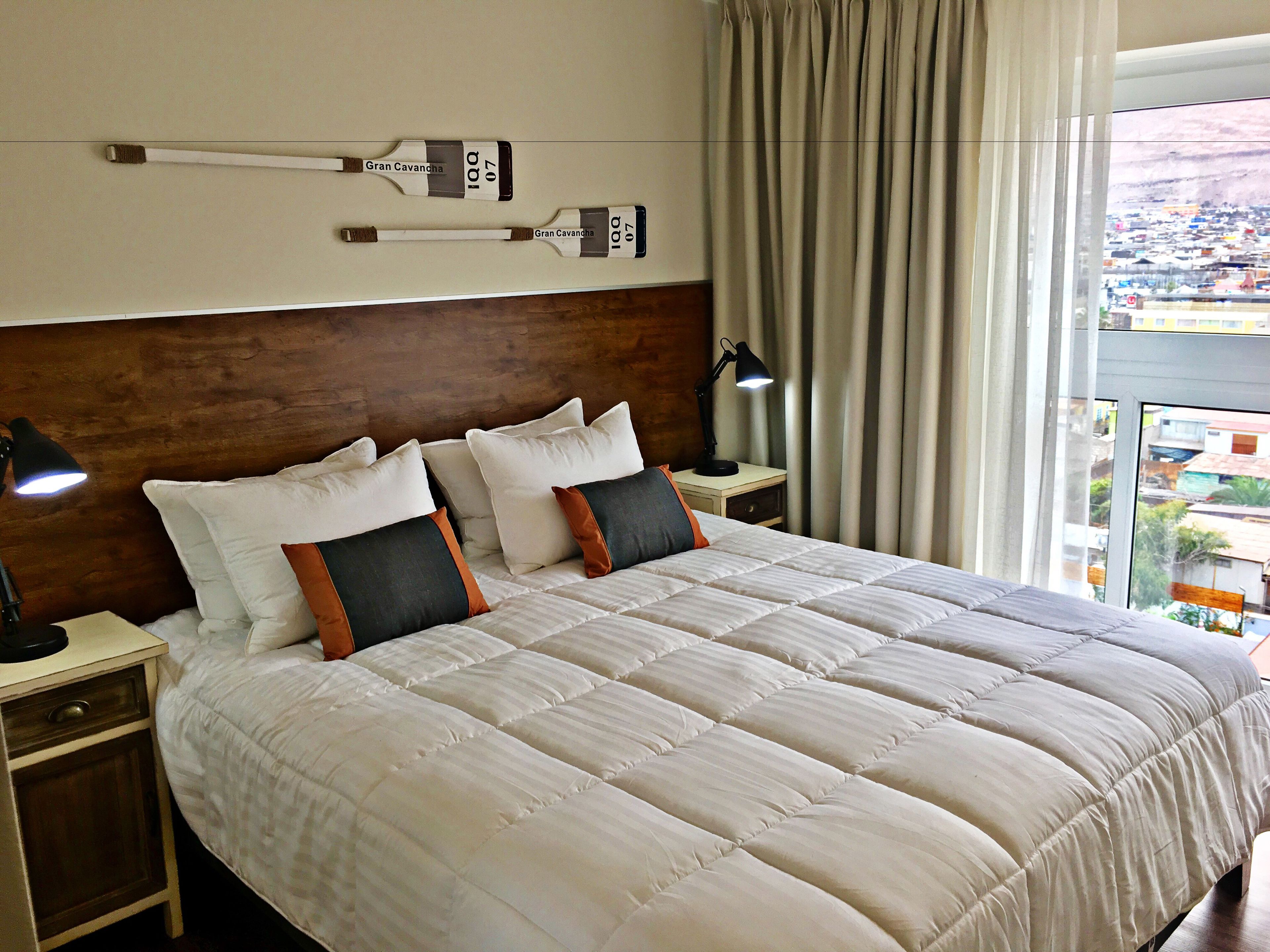 Quarto de hotel com uma cama grande, duas mesas de cabeceira com abajures, uma cabeceira de madeira e vista para a cidade pela janela.
