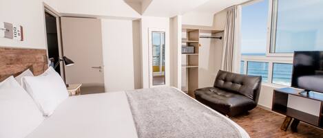Habitación de hotel con una cama grande, un sillón de cuero, un televisor y vista al océano.