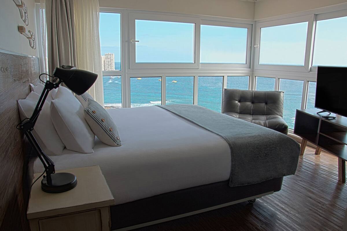 Habitación de hotel con una cama grande, una silla, un escritorio con lámpara, televisión y vista al océano.