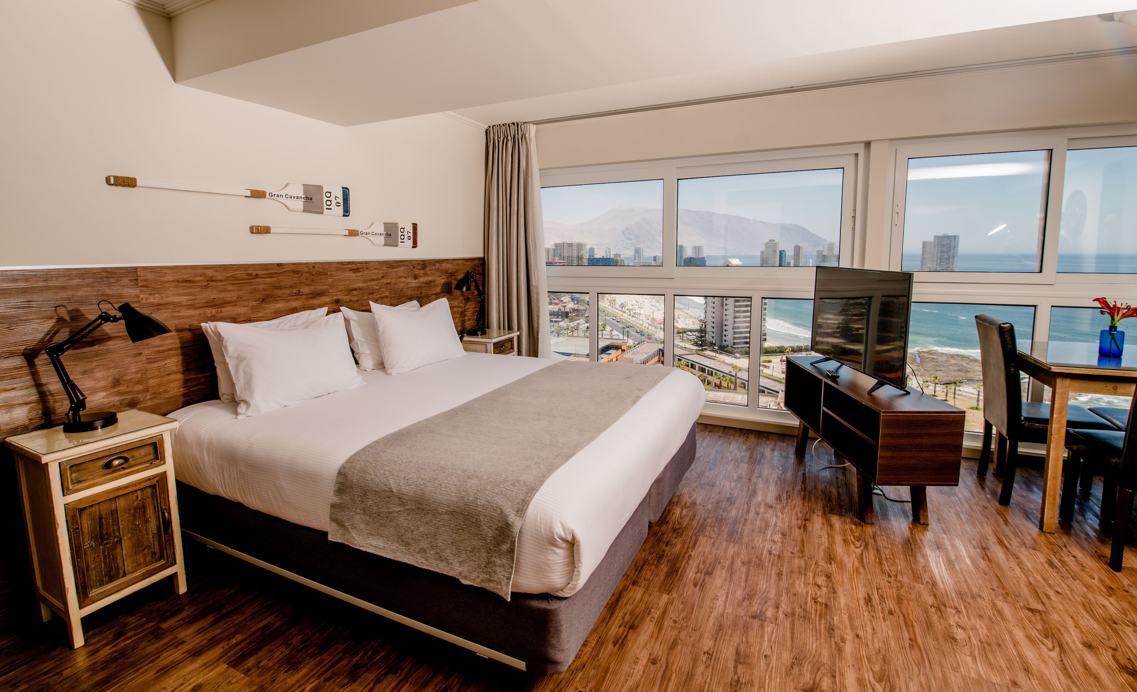 Habitación de hotel con una cama grande, un escritorio con televisión, una mesita de noche de madera y vistas al océano y al paisaje urbano.