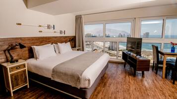 Habitación de hotel con una cama grande, un escritorio con televisión, una mesita de noche de madera y vistas al océano y al paisaje urbano.