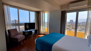 Habitación de hotel con cama, televisor, sillón y ventanas amplias con vista a la ciudad.