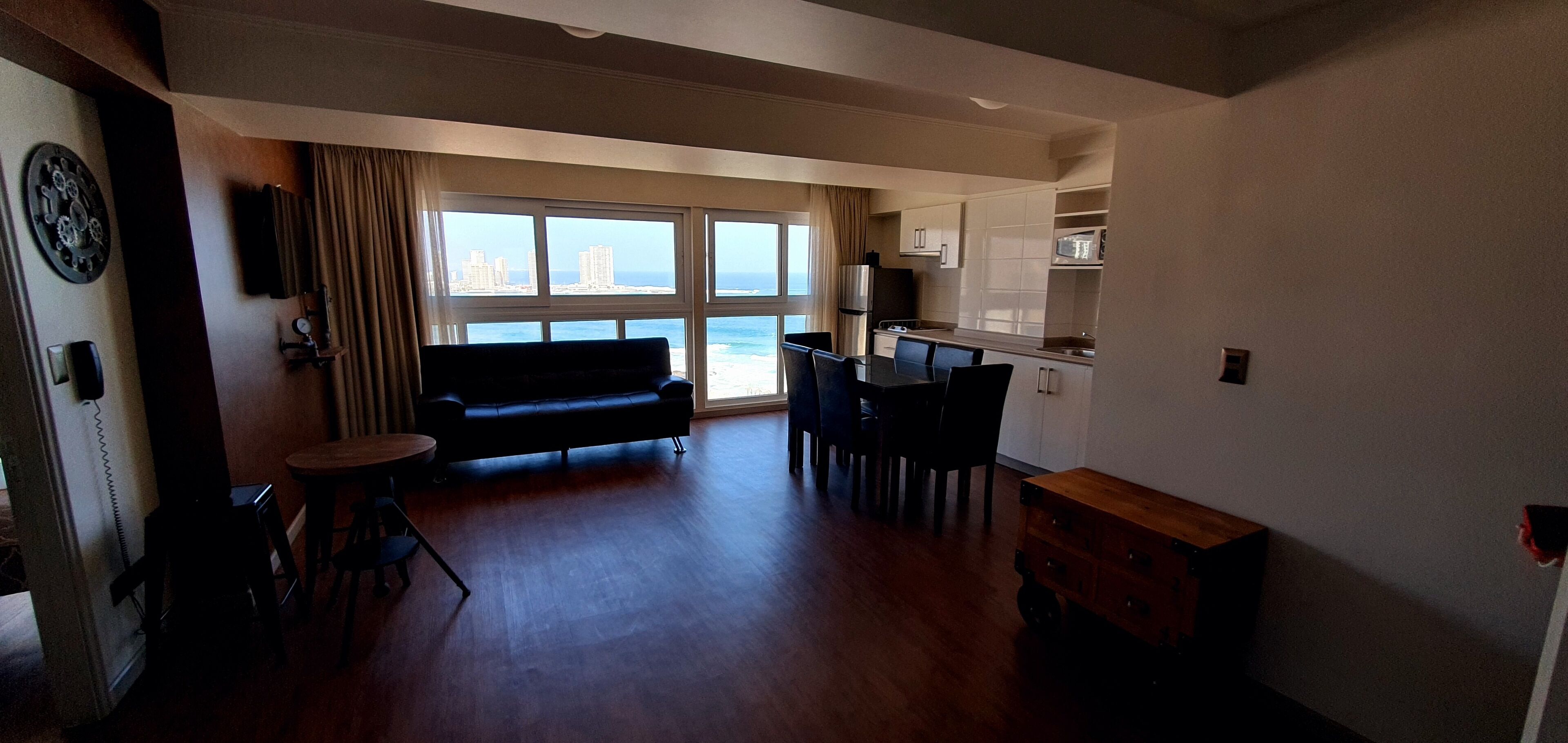 A living room with a sofa, dining table, and chairs, a kitchen area, and a large window with a view of buildings and the sea.