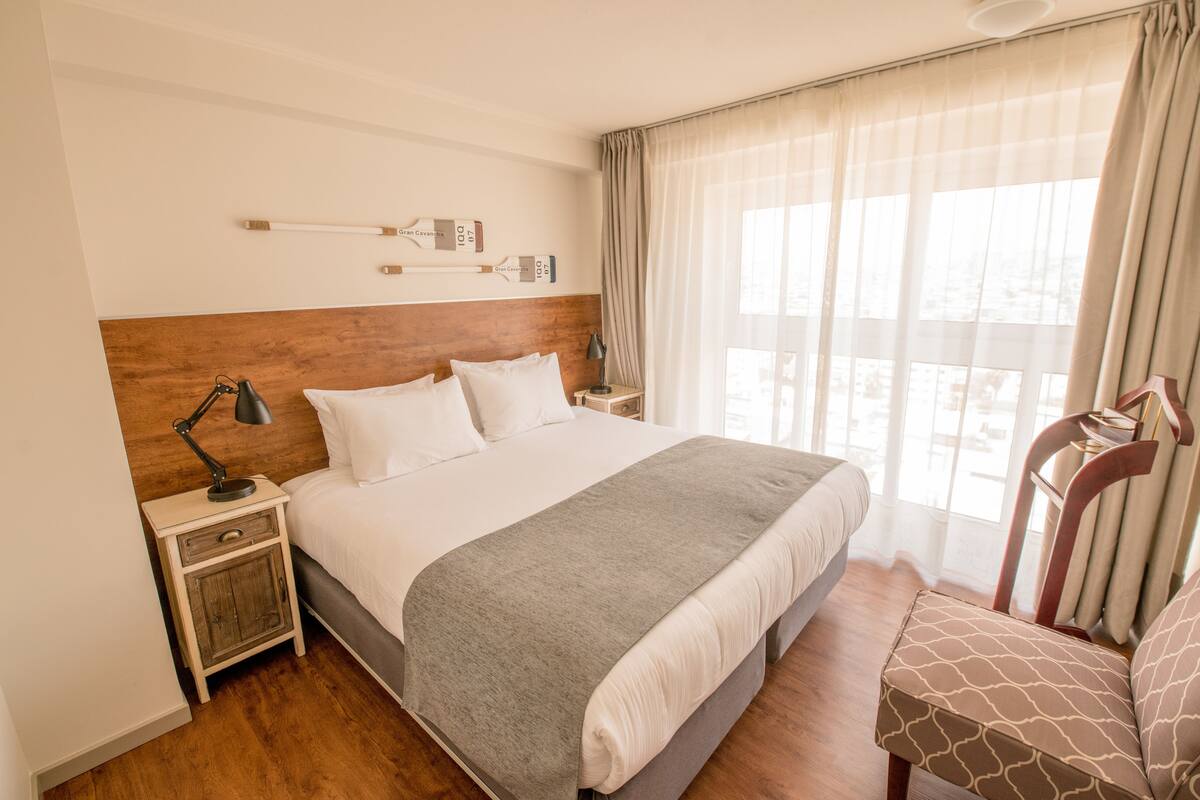 Una habitación de hotel ordenada con una cama grande, una mesita de noche con lámpara, una silla y una ventana con cortinas.