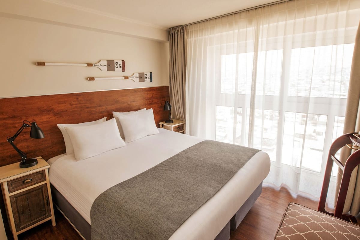 Habitación de hotel con una cama grande, cabecera de madera, mesita de noche con lámpara, pequeña mesa con lámpara y vista de la ciudad a través de cortinas traslúcidas.